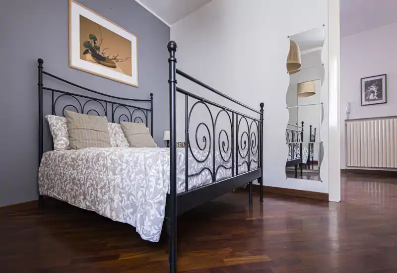 Bright, stylish bedroom with an ornate black metal bedframe, patterned bedcover and decorative cushions. Warm wooden floors and a full-length mirror create depth and a cozy atmosphere.