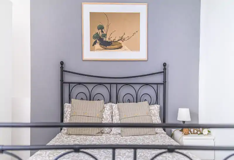 Bright, well-styled bedroom with a metal headboard, patterned bedding and coordinated cushions; bedside table with lamp and framed artwork above the bed.