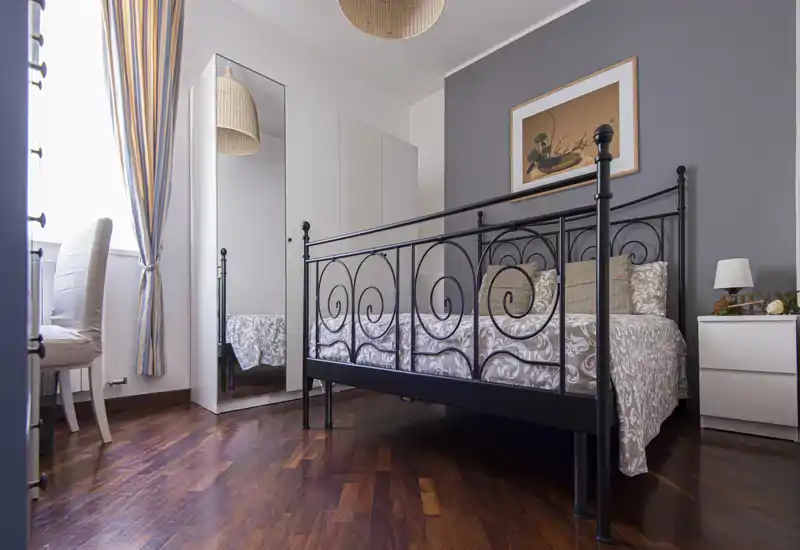 Bright, stylish bedroom with an ornate metal bed frame, patterned bedding and warm wooden floors. A full-length mirror, bedside table and natural light from the window complete the inviting layout.