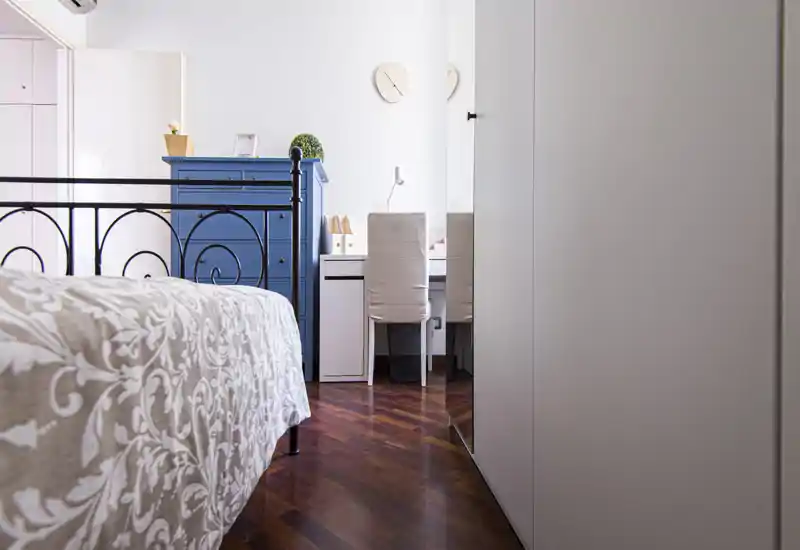 Bright, well-styled bedroom with patterned bedding, a metal bed frame and a blue chest of drawers. A small desk and chair are visible, offering a cozy workspace in the room.