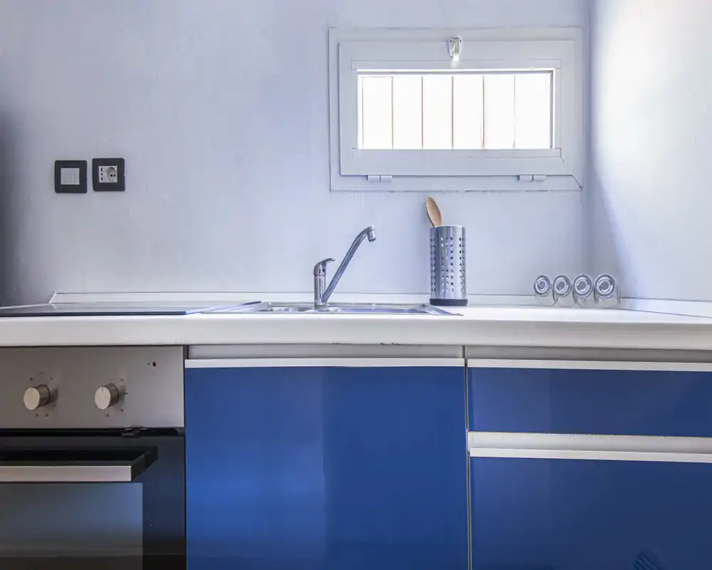 Modern compact kitchen with blue cabinetry, stainless sink and small window providing natural light — clean and well organized for rental photos.