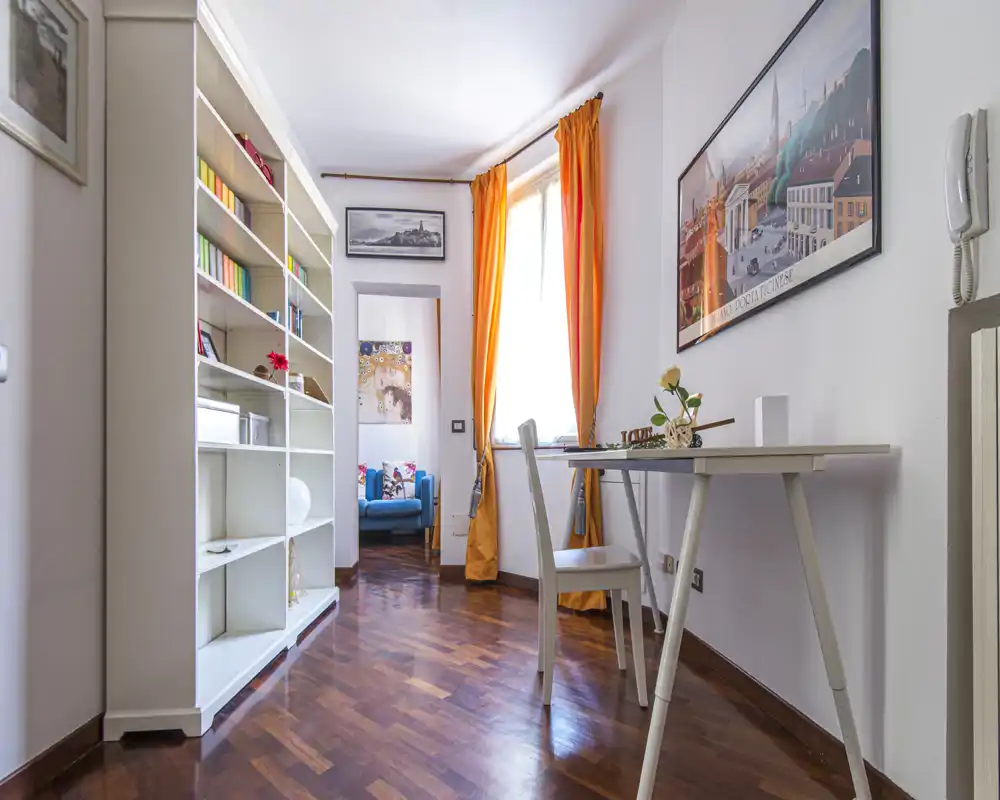 Bright, stylish living space with a slim desk, shelving unit and a cozy armchair visible through the doorway; warm orange curtains and wooden floors create a welcoming atmosphere.