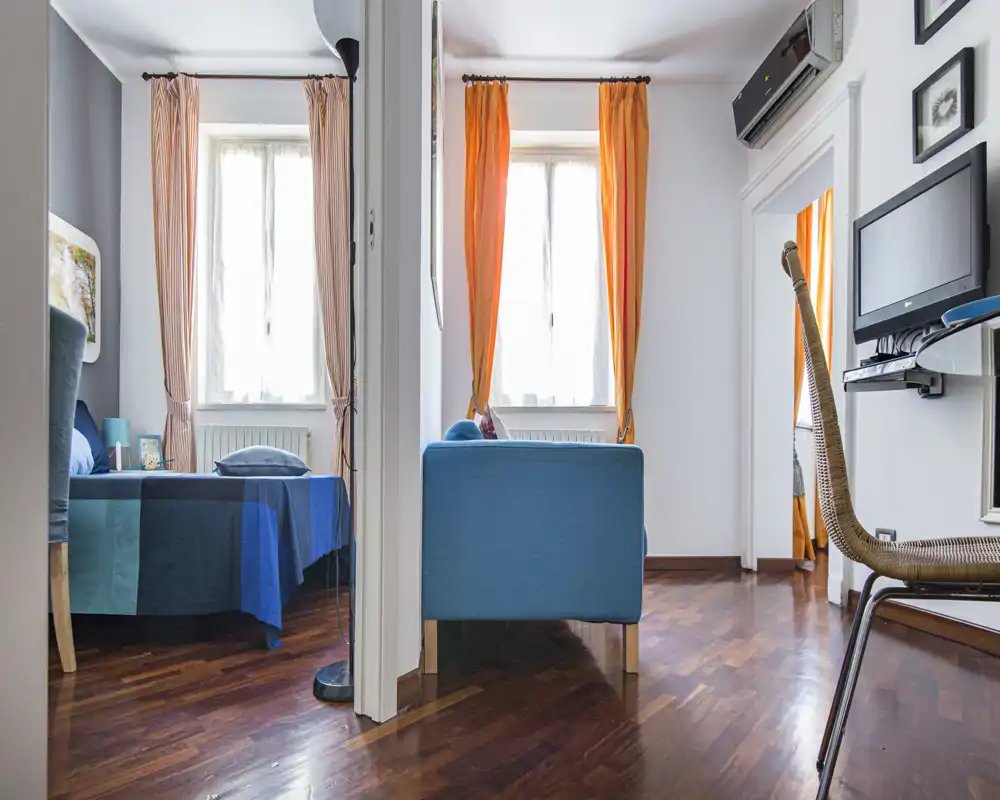 Bright, well-styled bedroom area with a visible double bed dressed in blue linens, warm orange curtains and a small adjacent sitting corner — ideal for a cozy city stay.
