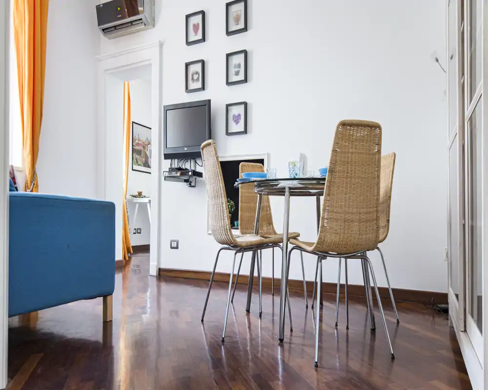 Bright, modern living area with a small dining table and wicker chairs on polished wooden floors; tasteful wall art and a blue sofa create a cozy, well-lit space ideal for short-term rentals.