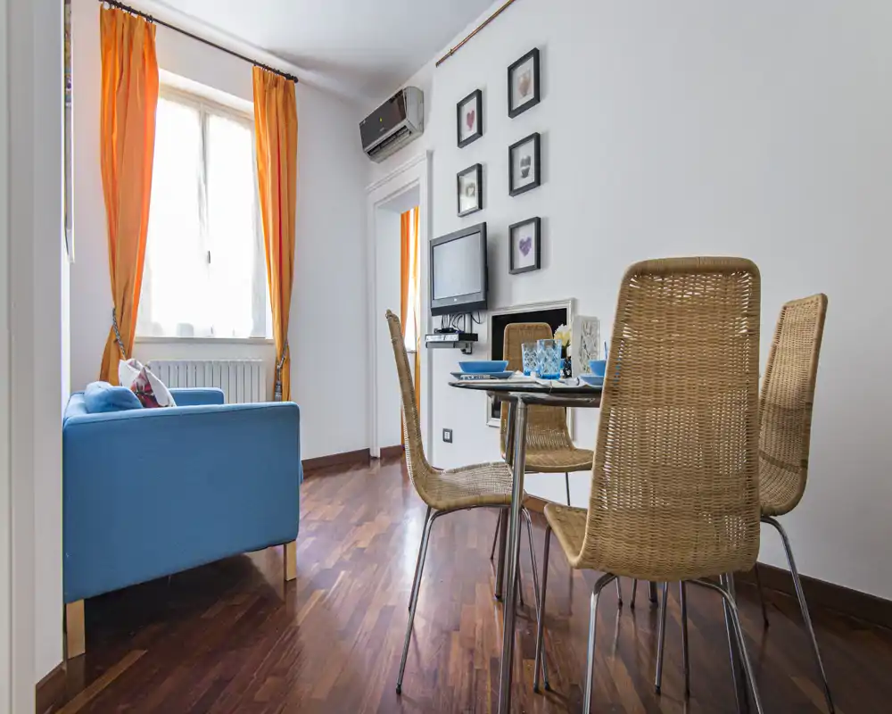 Bright, well-staged living area with a blue sofa, dining table and wicker chairs; warm orange curtains and wall art create a cozy atmosphere.