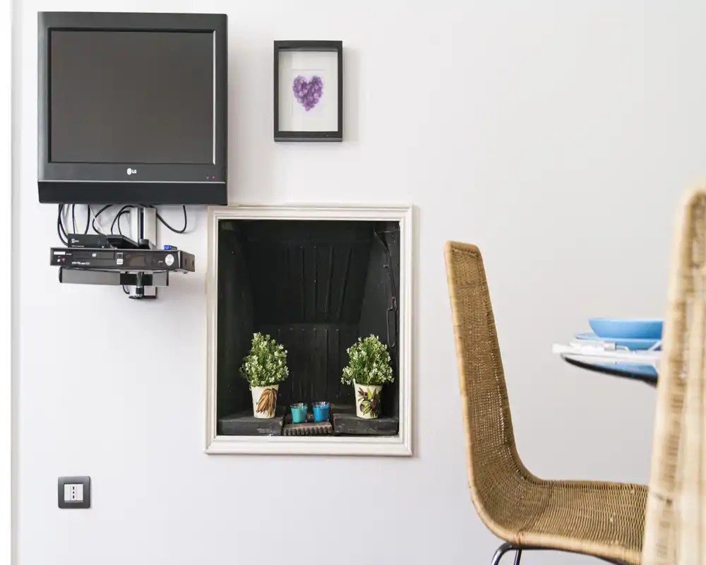 Bright, minimalist living area corner with a wall-mounted TV, small decorative niche with potted plants, and a woven chair next to a dining surface.