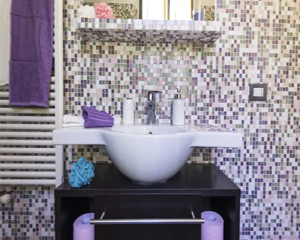 Contemporary bathroom sink with a rounded white basin set on a dark vanity, colorful mosaic wall tiles and neat accessories for a fresh, modern feel.
