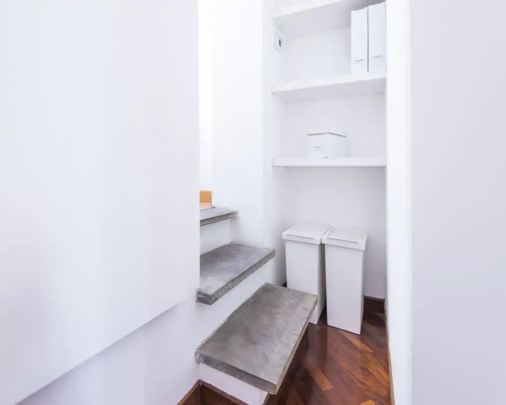 Small entrance/landing with stone steps, built-in shelving and two slim waste bins; bright white walls and polished wood floor create a clean, minimal look.