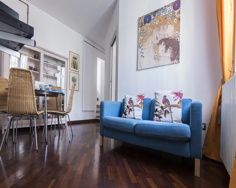 Bright and stylish living area with a compact blue sofa, decorative bird cushions, and a dining table set nearby. Warm wooden floors and colorful artwork create a cozy, well-styled apartment corner.