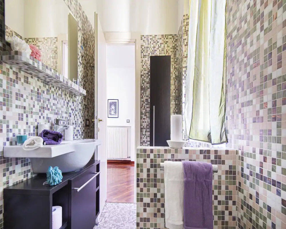 Bright, modern bathroom with mosaic tiles, a wall-mounted sink, towel rail and a window with a light curtain letting in natural light.