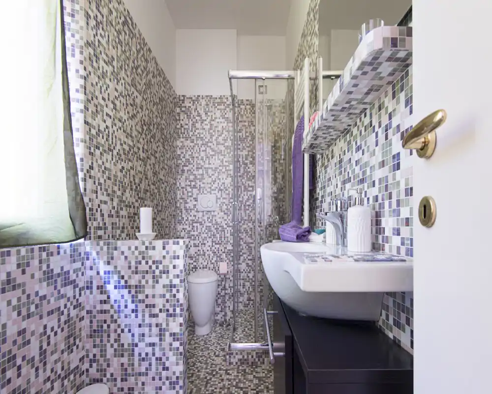 Bright, modern bathroom with mosaic tiles covering walls and floor, a glass shower enclosure, wall-mounted sink and toilet. Clean, compact layout with purple towel accents and good natural light.