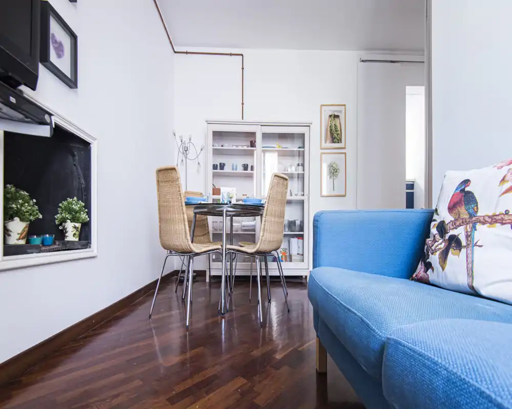 Bright, well-styled living area with a blue sofa, decorative cushion and a small dining table; polished wooden floors and framed wall art create a cozy, modern atmosphere.