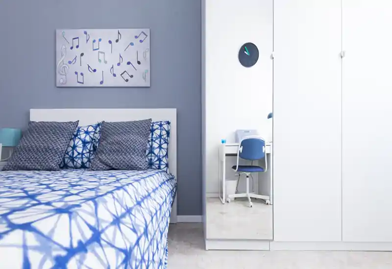 Bright, tidy bedroom with blue-and-white linens, decorative pillows and a mirrored wardrobe that adds depth; compact desk is visible through the mirror.