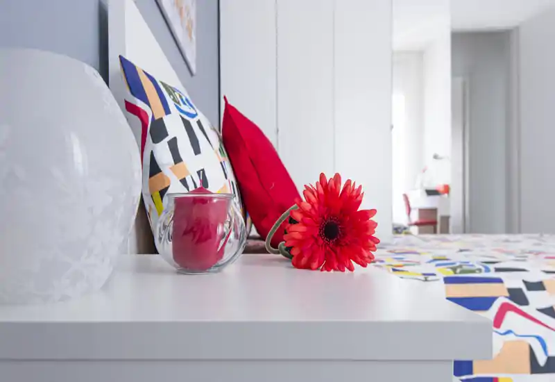 Bright, modern bedroom detail with colorful pillows, patterned bedding and a decorative flower on the bedside table — a fresh, welcoming composition ideal for rental photos.