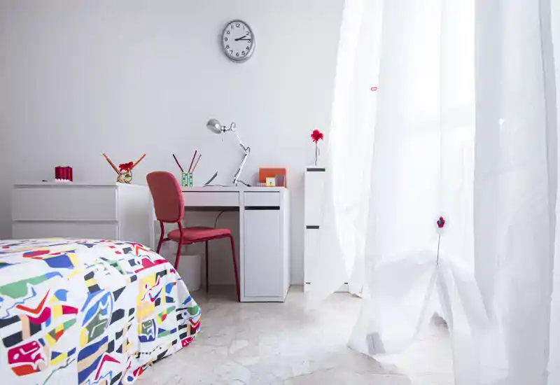 Bright, minimal bedroom with a colorful bedspread, white desk and a red chair beside a large window letting in abundant daylight.