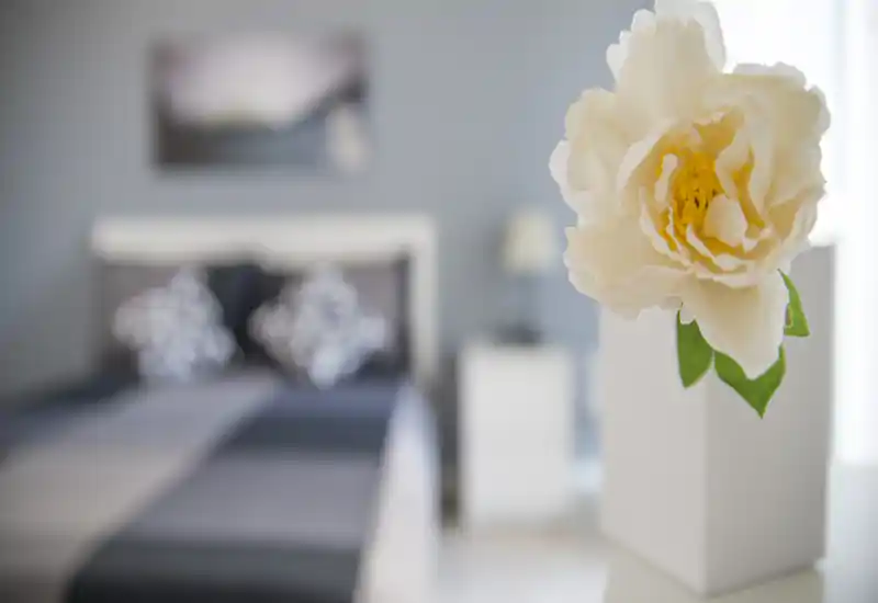 Chambre élégante avec lit légèrement flou et coussins décoratifs ; ambiance lumineuse et moderne avec un accent floral au premier plan.