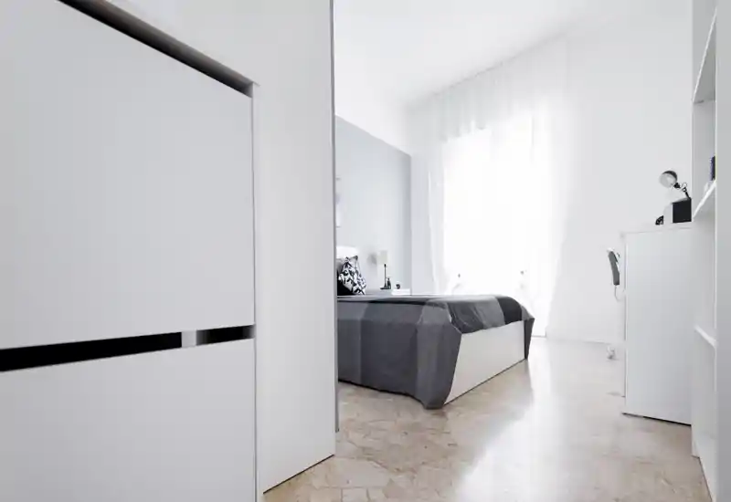 Chambre minimaliste et lumineuse avec un lit double en linge gris, grande fenêtre aux voilages légers et mobilier blanc moderne — espace épuré et aéré, parfait pour se reposer.