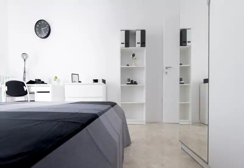 Chambre minimaliste et lumineuse avec un lit double bien fait, des rangements blancs et une grande glace reflétant la pièce. Lignes épurées et lumière naturelle pour un espace de nuit moderne et apaisant, idéal pour une annonce.