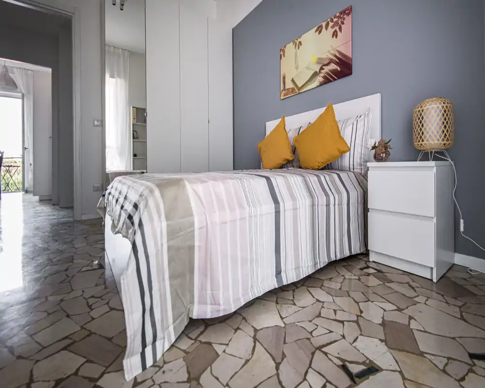 Chambre lumineuse et bien décorée avec un lit double à rayures, coussins moutarde, table de chevet et lampe, et un mur gris apportant une ambiance apaisante.