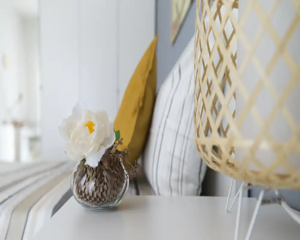 Gros plan d'une table de chevet décorée avec un vase, des coussins et une lampe tressée; lumière naturelle douce et textiles pour une ambiance chaleureuse.