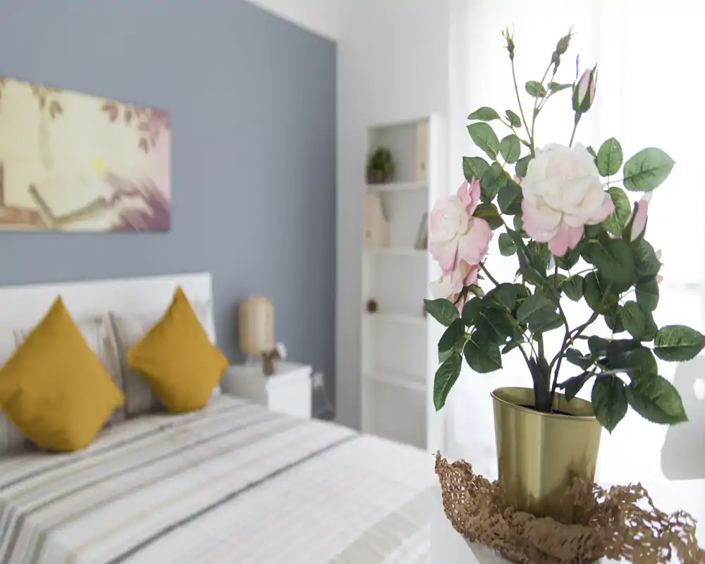 Chambre lumineuse et stylée avec un lit bien fait, des coussins moutarde, une lampe de chevet et une décoration murale; lumière naturelle agréable et décor moderne soigné.