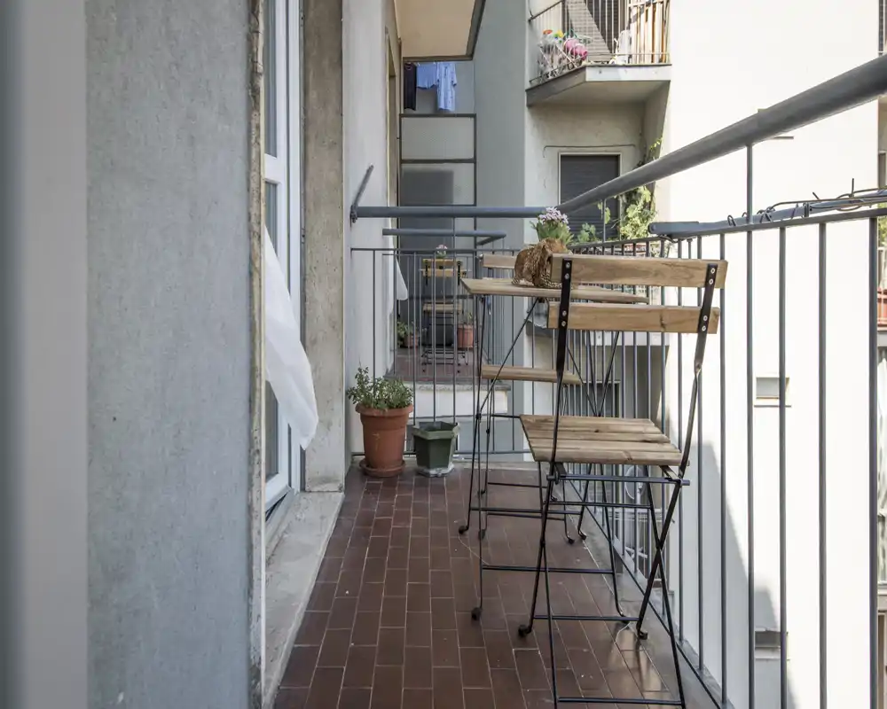 Balcon étroit ensoleillé avec une petite table bistro en bois et deux chaises pliantes, sol en terre cuite et plantes en pots — idéal pour un café le matin.