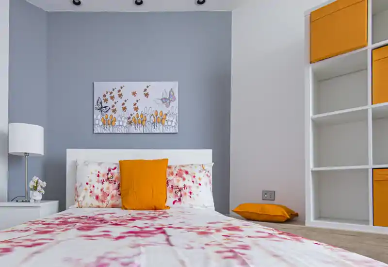 Camera da letto moderna e luminosa con piumone floreale e vivaci tocchi arancioni — comodo letto matrimoniale, lampada da comodino e scaffalatura integrata.