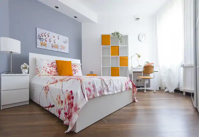 Camera da letto moderna e luminosa con letto bianco, biancheria floreale, cuscini arancioni e una scrivania vicino alla finestra. Pavimento in legno e scaffalatura minimal conferiscono un'atmosfera pulita e accogliente.