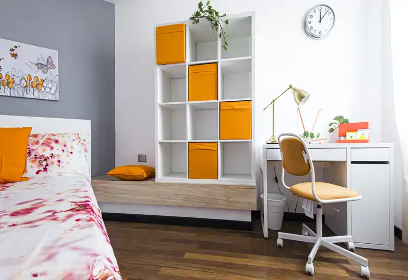 Camera da letto luminosa e moderna con struttura del letto bianca, biancheria floreale e particolari arancioni; mobile a cubi con contenitori arancioni e scrivania vicino alla finestra.