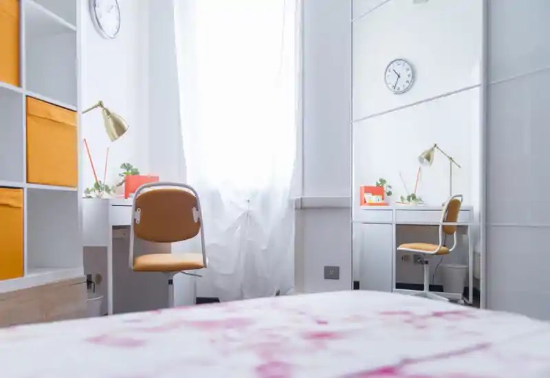 Dormitorio luminoso y ordenado con cama en primer plano, escritorio y silla junto a la ventana iluminada y armario con espejo que aporta más profundidad.