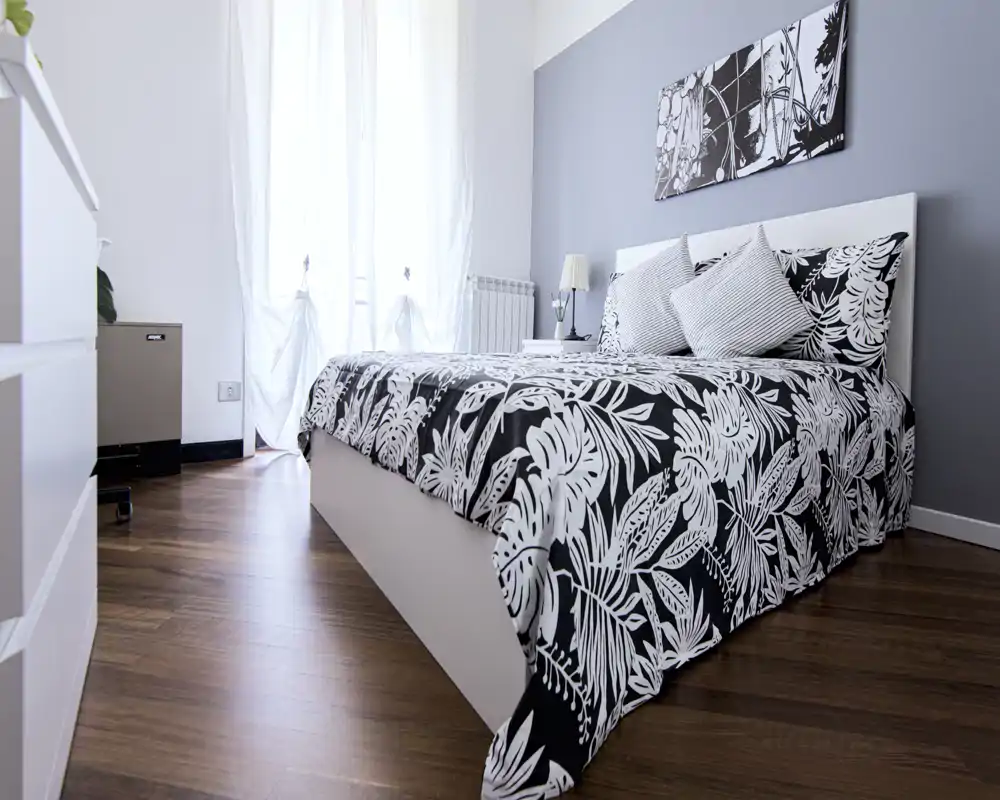 Dormitorio moderno y luminoso con colcha estampada en blanco y negro, cojines decorativos y cabecero minimalista. La luz natural entra por una ventana alta, destacando el suelo de madera y un cuadro sencillo.