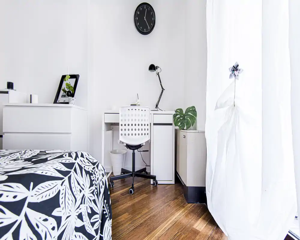 Dormitorio luminoso y minimalista con colcha estampada, escritorio y silla blancos compactos, y luz natural de una gran ventana—ideal para estudiante o alquiler individual.