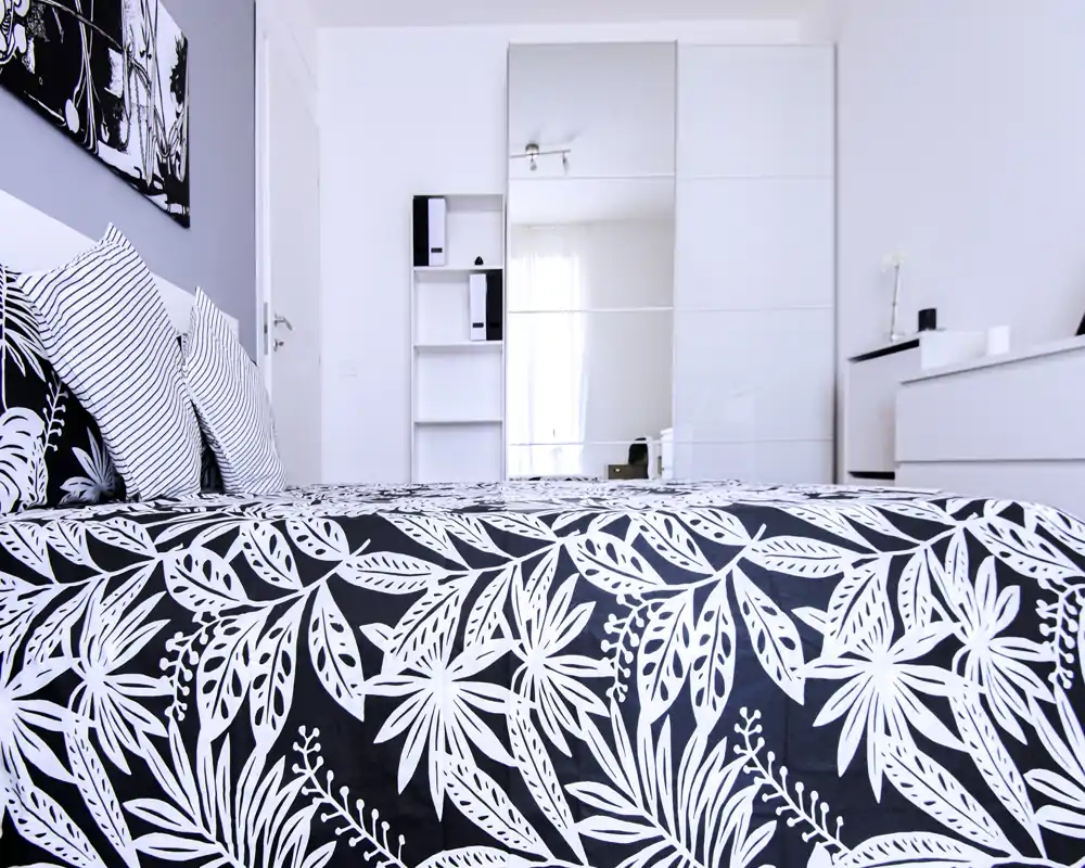 Dormitorio moderno y luminoso con colcha de estampado llamativo, cojines a rayas y armario con espejo que refleja luz natural; decoración minimalista y estilosa, ideal para un anuncio de alquiler.