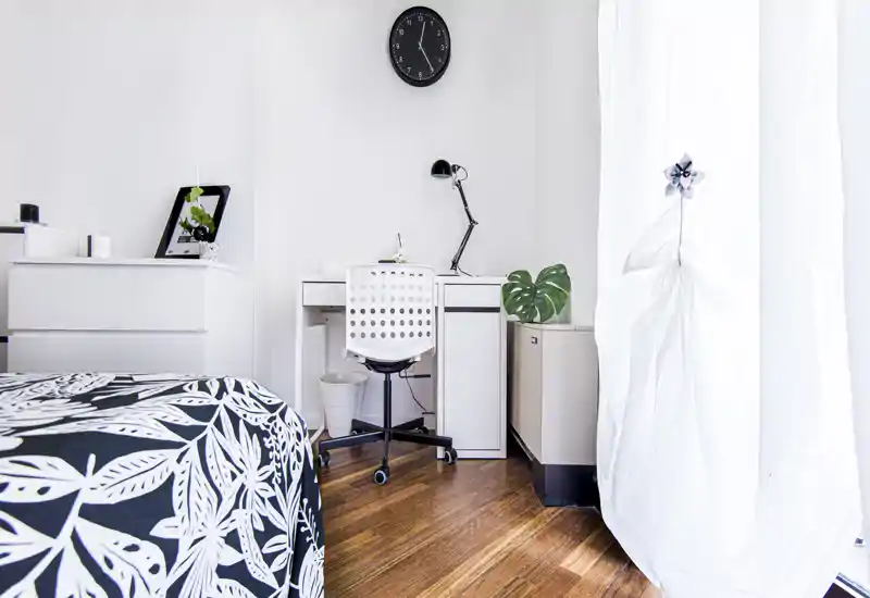 Camera da letto luminosa e minimal con copriletto a motivi, una scrivania bianca compatta e sedia, e luce naturale da una grande finestra—ideale per monolocali o studenti.