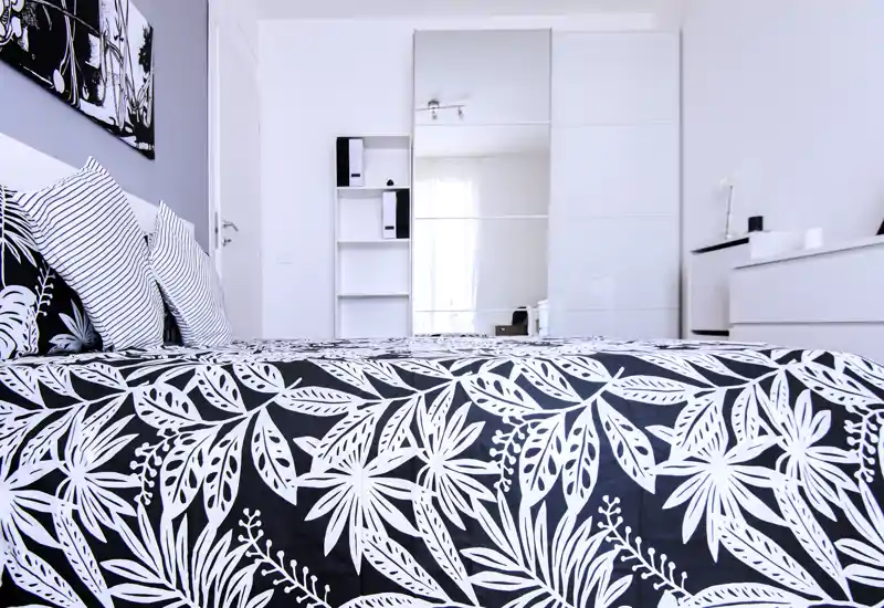 Camera da letto moderna e luminosa con copriletto a motivo deciso, cuscini a righe e guardaroba con ante a specchio che riflette luce naturale; arredamento essenziale e di stile, perfetto per un annuncio.