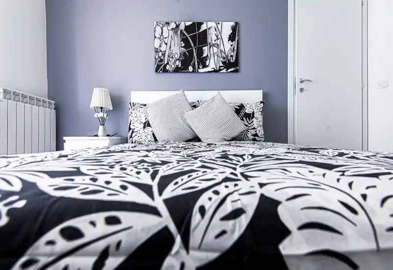 Camera da letto luminosa e di stile con un grande letto a fantasia in primo piano, cuscini coordinati, comodino e lampada su una parete grigio tenue — ideale per annunci di affitto.