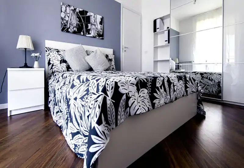 Camera da letto luminosa e di design con letto matrimoniale completo, copriletto botanico bianco e nero, comodino con lampada e armadio con ante a specchio che riflettono la luce naturale.