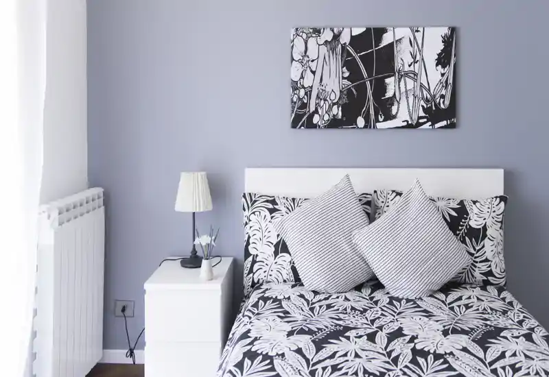 Camera da letto luminosa e di stile con copriletto a fantasia, due cuscini decorativi e un comodino minimal. La luce naturale da sinistra illumina la parete grigio chiaro e l'opera monocromatica sopra la testata.