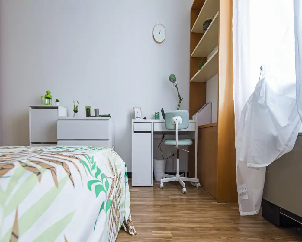 Bright, modern bedroom corner with a patterned bed in the foreground and a tidy study desk with shelving by the window. Natural light creates a welcoming atmosphere and the room features clean, minimal decor.