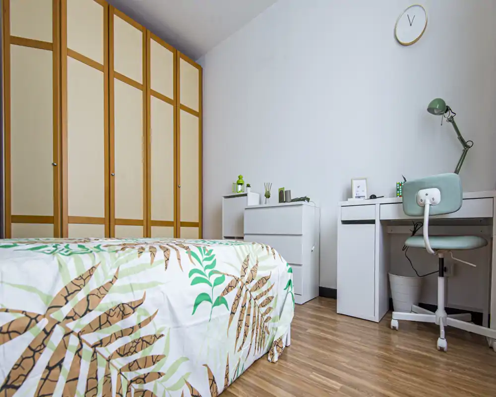 Bright, tidy bedroom with a patterned bedspread, large built-in wardrobe and a compact desk area — ideal for a student or single occupant.