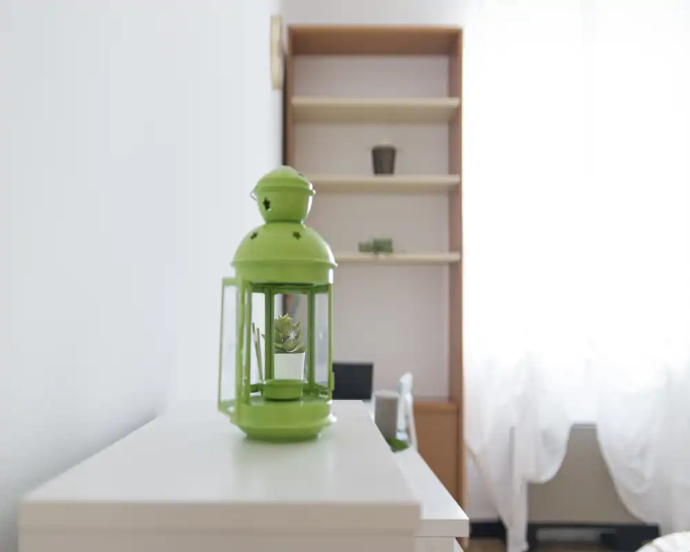 A close-up decorative shot of a green lantern placed on a white dresser or shelf; a tall shelving unit and a bright window with sheer curtains are visible in soft focus behind it.