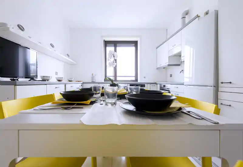 Cucina moderna e luminosa con tavolo apparecchiato per quattro, sedie gialle, stoviglie nere e luce naturale dalla finestra centrale.