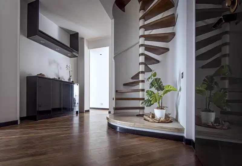 Ingresso luminoso e moderno con scala a chiocciola in legno, pavimenti in parquet lucido e un elegante mobile nero; una pianta e decor minimal rendono l’ambiente accogliente.