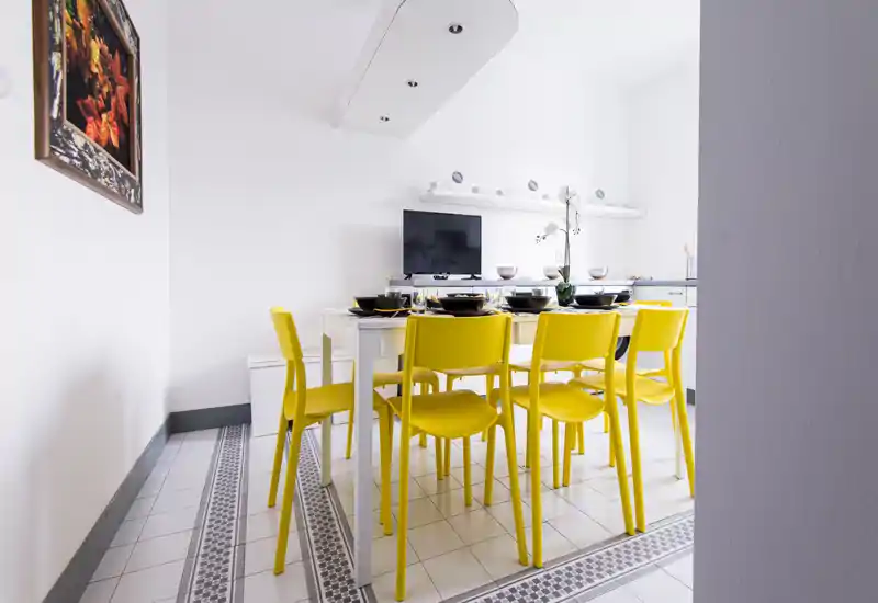 Zona pranzo luminosa con tavolo bianco e sei vivaci sedie gialle, apparecchiata con stoviglie e un semplice centro tavola con orchidea — ambiente moderno e allegro ideale per i pasti.