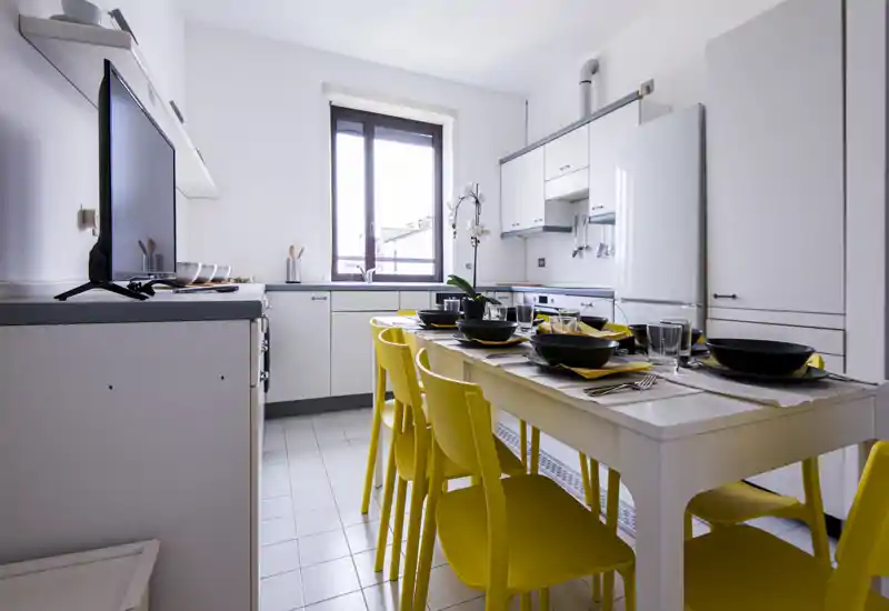 Cucina moderna e luminosa con mobili bianchi, elettrodomestici integrati e tavolo apparecchiato con sedie gialle vivaci — perfetta per annunci fotografici e pasti in compagnia.
