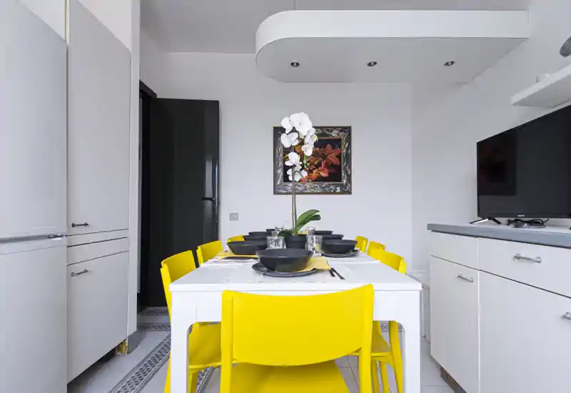 Area pranzo moderna e luminosa con tavolo bianco per sei e sedie gialle vivaci; un'orchidea centrale e stoviglie nere creano un elegante contrasto.