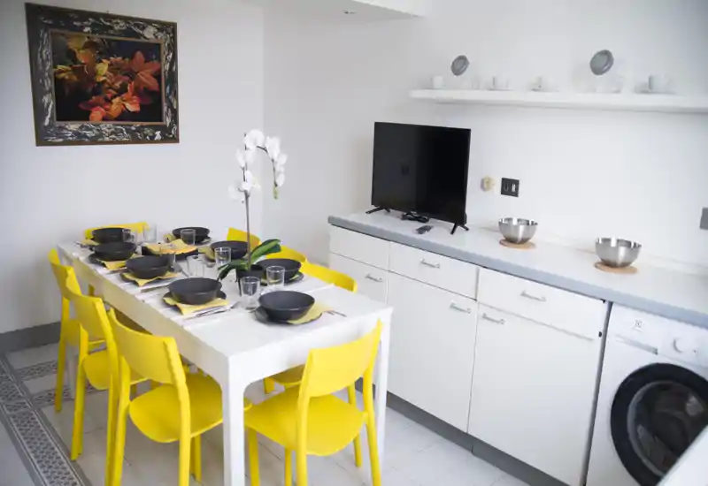 Zona pranzo moderna e luminosa con tavolo bianco per sei persone e sedie gialle vivaci; i coperti ordinati e un'orchidea centrale creano un'atmosfera accogliente.
