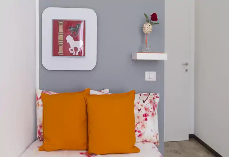 Angolo di camera piccolo e curato con letto visibile con biancheria floreale e due cuscini arancioni vivaci, parete grigia, quadro decorativo e una mensola a scomparsa con un vaso.