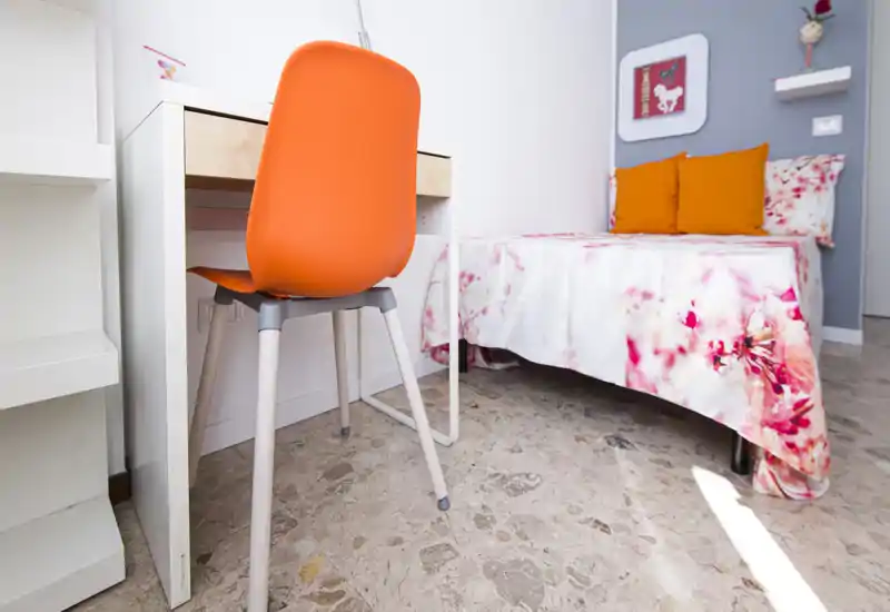 Accogliente camera singola con copriletto floreale e cuscini arancioni vivaci; una scrivania compatta e una sedia arancione creano un angolo studio allegro. La luce naturale illumina la stanza mettendo in risalto l'arredamento colorato.