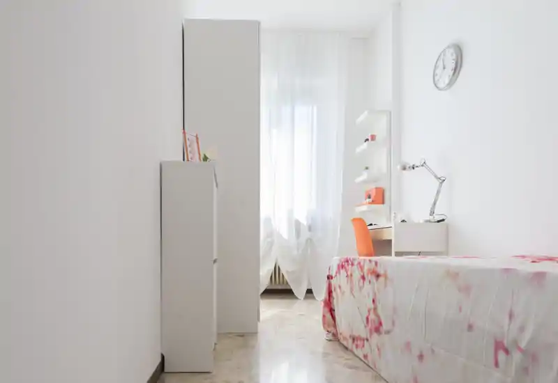 Camera da letto singola luminosa e minimale con copriletto fantasia, una scrivania compatta e una sedia arancione vicino alla finestra. Pareti bianche, tende leggere e mensole semplici creano un’atmosfera fresca e ariosa, ideale per una persona.